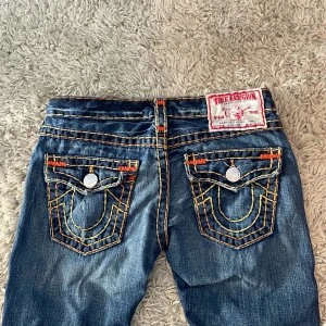 Blå Ture religion jeans - Snygga blå jeans från True Religion med iögonfallande kontrastsömmar i orange och gult. Jeansen är low wasted. Byxorna har en bootcut-stil och är dekorerade med knappar och broderade detaljer på bakfickorna. Perfekta för en cool och avslappnad look. 🫶🫶