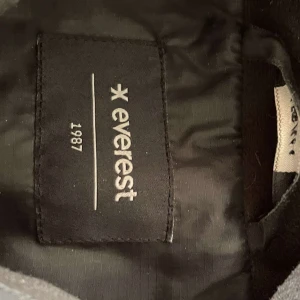 Svart dunväst från Everest - Säljer en svart dunväst från Everest med dragkedja framtill. Västen har en quiltad design och praktiska fickor på sidorna. Har även en innerficka på vänster sida av västen. Perfekt för kyligare dagar när du vill hålla värmen utan att kompromissa med stilen.