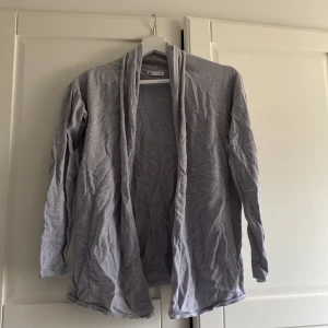 Grå kofta - Säljer en stilren grå kofta med lång ärm. Perfekt för lager-på-lager under kyligare dagar. Koftan har en öppen design utan knappar eller dragkedja, vilket ger en avslappnad look.