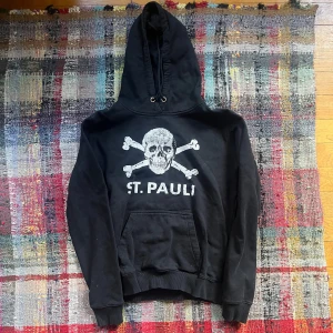 Svart hoodie från St. Pauli - Säljer en svart hoodie från St. Pauli med ett stort dödskalletryck och korsade benknotor på framsidan. Hoodien har en klassisk känguruficka och justerbar huva med dragsko. Perfekt för en avslappnad stil.