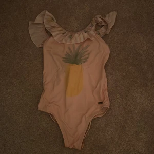 Rosa baddräkt med volang från Patachou - Söt rosa baddräkt från Patachou med volangdetaljer runt halsen och en ananastryck på framsidan. Baksidan har en fin snörning och små rosetter. Perfekt för sommarens badäventyr! Den har dock fått lite fläckar inuti, men det är inget som syns utanpå och går nog bort om man försöker. Hittar ingen storlek men funkar för barn mellan 8-10 år.