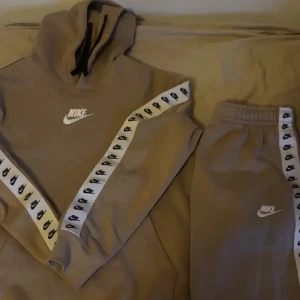 Beige hoodie från Nike - Säljer en beige hoodie från Nike med vit logga på bröstet och svarta detaljer. Ärmarna har en vit rand med Nike-loggor längs sidorna. Perfekt för en avslappnad stil.