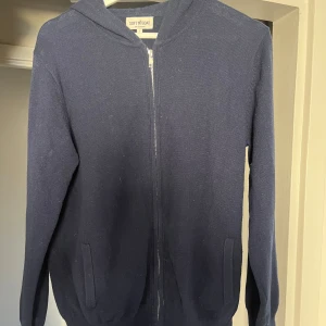 Mörkblå hoodie från Soft Goat - Säljer en stilren mörkblå hoodie från Soft Goat i 100% kashmir i herr modellen men passar båda. Tröjan har dragkedja framtill och långa ärmar, perfekt för kyliga dagar. Den är både mjuk och bekväm, med en klassisk design som passar till det mesta.