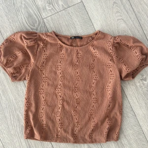 Brun topp från Zara - Säljer en söt brun topp från Zara med korta puffärmar och ett vågigt hålmönster. Perfekt för en stilren look. Toppen har en rund halsringning och är gjord i ett mjukt material.