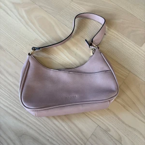Rosa axelväska från Don Donna - Snygg rosa axelväska från Don Donna i syntetmaterial. Väskan har en praktisk dragkedja och en stilren design som passar perfekt för alla tillfällen. Dennöe aldrig använd så har därför ett jättebra skick, original pris är 1000kr