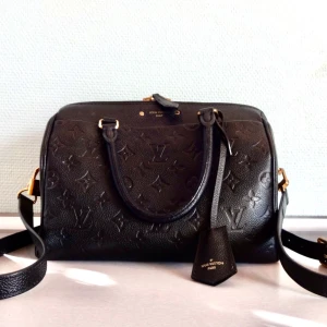 Louis Vuitton Speedy Bandouliere 25 in Black Empreinte Leather with Gold Hardware - The bag has one slip pocket on the outside, two slip pockets, and one zipper pocket on the inside. The lining of the bag is textile.   The size of this bag is 25 x 19 x 15 cm.    The things you need to consider before buying this bag :  -The hardware of the bag begins to fade. -There are some dents on the bag. -The corner of the bag is painted.  -One of the handle’s glazing begins to get cracked. -There are some small stains on the inside of the bag.   The bag will be delivered without dustbag.