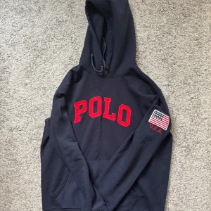 Mörkblå hoodie från Polo Ralph Lauren - Säljer en snygg mörkblå hoodie från Polo Ralph Lauren med röd text på bröstet och en amerikansk flagga på ärmen. Perfekt för en casual look. Den har en klassisk känguruficka och justerbar huva.