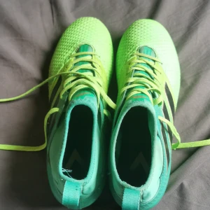 Gröna fotbollsskor från Adidas - Snygga gröna fotbollsskor från Adidas med snörning och en mönstrad ovandel. Skorna har dobbar för bra grepp på planen och en sportig design med svarta detaljer.