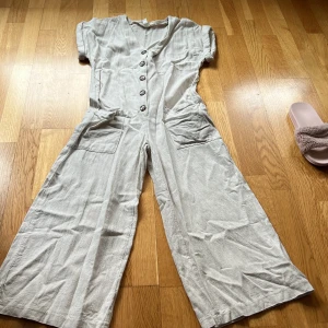 Beige linnen jumpsuit från H&M - Säljer en stilren beige jumpsuit från H&M med knappar framtill och korta ärmar. Den har praktiska fickor och ett knytband i midjan för en justerbar passform. Perfekt för en avslappnad look! 
