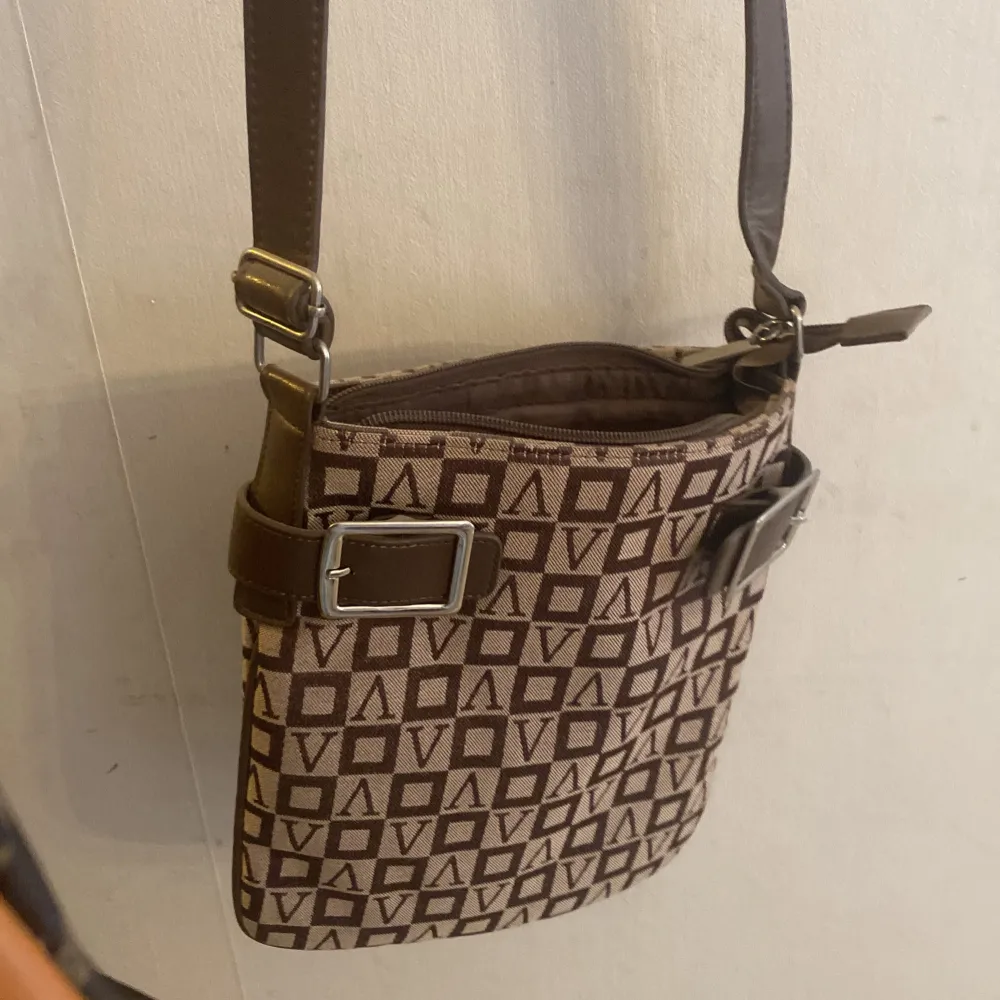 Snygg brun axelväska med geometriskt mönster och justerbar axelrem. Väskan har en praktisk dragkedja och dekorativa spännen på sidorna. Perfekt för att bära dina dagliga nödvändigheter med stil.. Laukut & Käsilaukut.