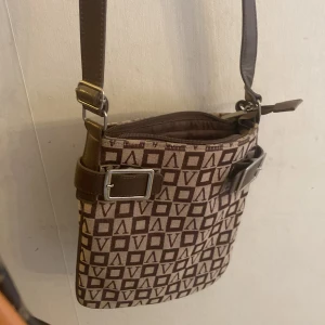 Brun mönstrad axelväska - Snygg brun axelväska med geometriskt mönster och justerbar axelrem. Väskan har en praktisk dragkedja och dekorativa spännen på sidorna. Perfekt för att bära dina dagliga nödvändigheter med stil.