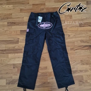 Svarta Corteiz Cargobyxor - Säljer helt nya Corteiz Guerillaz Cargo Pants i svart med rosa detaljer, originaletiketter kvar. Storlek: L (passar mer som M). Material: Slitstarkt ripstop-tyg, bekvämt. Design: Broderade & tryckta detaljer, cargofickor med lock, justerbara band i midja & benslut. Perfekt för dig som gillar exklusiv streetwear! Limited release – svåra att få tag på! 📦 Snabb leverans – skickas samma dag! 📦 Kontakta mig vid intresse eller funderingar!📌