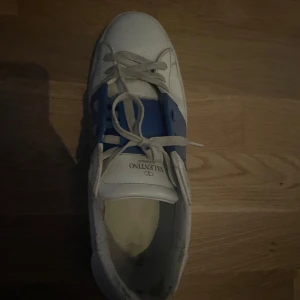 Valentino sneakers med blå detalj - Snygga vita Valentino sneakers med en iögonfallande blå detalj över snörningen. Skorna har en klassisk rund tå och är tillverkade i skinn. Perfekta för en stilren look.