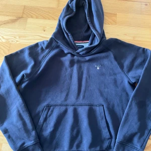 Mörkblå hoodie från Gant - Säljer en snygg mörkblå hoodie från Gant. Den har en klassisk design med en känguruficka framtill och en liten logga på bröstet. Perfekt för en avslappnad stil.