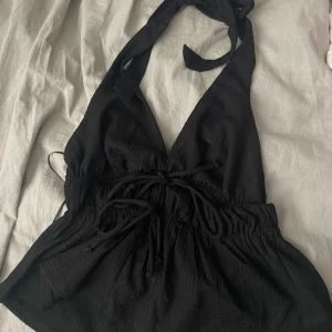 Svart halterneck topp - Snygg svart halterneck topp med knytband i nacken och midjan. Toppen har en djup urringning och elastisk midja för en bekväm passform. Perfekt för en stilren look.