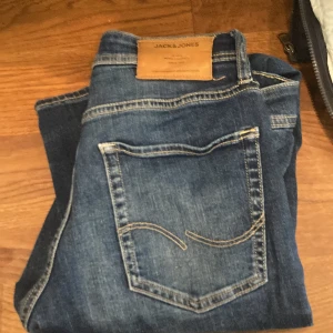Blå slim fit jeans från Jack & Jones - Snygga blå slim fit jeans från Jack & Jones, modell Slim Glenn. Perfekta för en stilren look med klassisk femficksdesign och knappgylf. Passar utmärkt för både vardag och mer uppklädda tillfällen.