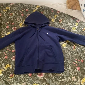 Polo ralph lauren zip hoodie - Riktigt snygg ralph lauren zip hoodie i en mörkblå färg som gör att den är enkel att styla, säljes pågrund av att jag inte använder den längre. Den enda defekten är att det hänger ut en tråd lite från snöret (se bild 3). Är osäker på äkthet därav priset
