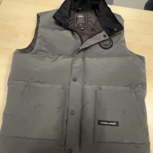 Canada Goose väst  - Säljer en stilren grå dunväst från Canada Goose med svart krage och knappar. Västen har två stora fickor framtill och ett broderat märke på bröstet. Perfekt för kyliga dagar.