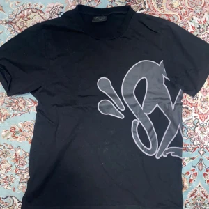 Svart Synaworld t-shirt  - Snygg svart t-shirt med ett stort, stilrent Synaworld teyck i grått som täcker både fram- och baksidan. Perfekt för en casual look.