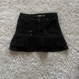 Svart kortkjol med volang och dragkedjor - En lågmidjad jeanskjol omsydd från ett par jeans Midjemått tvärsöver - 34 cm Längd - 33 cm