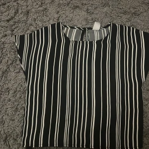 Svart och vit randig blus - Säljer en stilren svart och vit randig blus med korta ärmar. Blusen har en knappdetalj i nacken och är perfekt för en chic look. Passar bra till både jeans och kjol. 