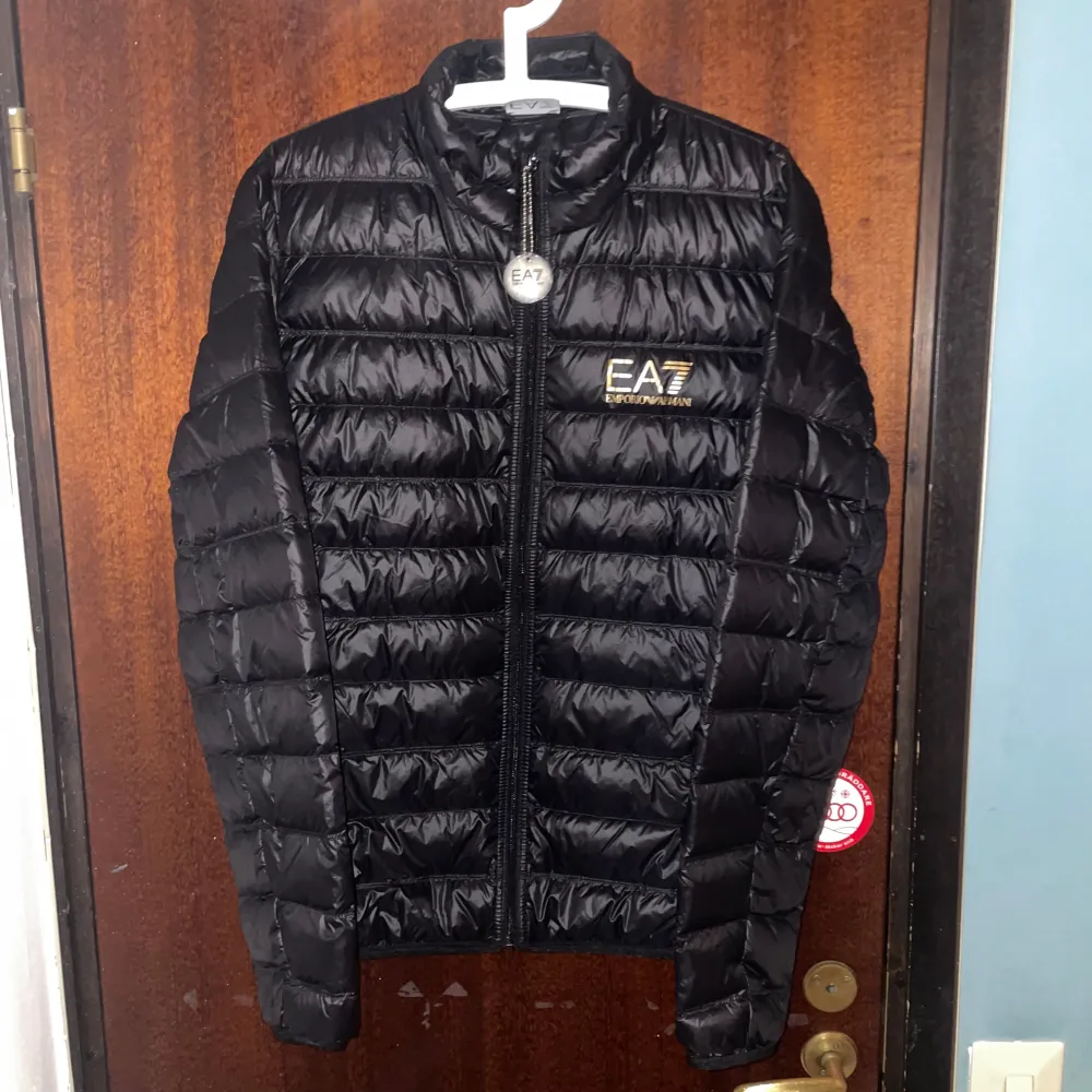 Snygg svart pufferjacka från EA7 Emporio Armani med dragkedja framtill. Jackan har en quiltad design och är perfekt för kyligare dagar. Den är lätt och smidig att packa ner i medföljande påse.. Takit.