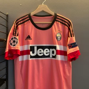 Rosa fotbollströja från Adidas - Säljer en rosa fotbollströja från Adidas med svarta och röda detaljer. Tröjan har korta ärmar och Juventus-logga på bröstet. Den har även texten 'Jeep' och 'Pogba 10' på ryggen. Perfekt för fotbollsfans! Helt ny med tagg i storlek M en replika 