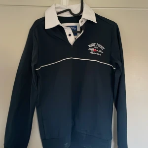Svart pikétröja med vit krage - Snygg svart pikétröja med vit krage och broderad text 'West Point Sailing Team' på bröstet. Tröjan har långa ärmar och en klassisk pikéknäppning framtill. Den är i storlek M men jag skulle uppskatta den som S.