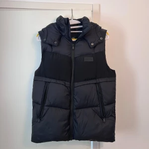 River Island väst - Snygg svart dunväst från River Island med dragkedja och huva. Perfekt för kyligare dagar och ger en stilren look. Västen har praktiska fickor med dragkedja och en diskret logotyp på bröstet. Västen har suttit i garderoben i snart ett år och har använts max 5 gånger. Vid frågor eller behov av fler bilder, är det bara att fråga. 