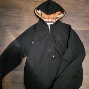 Svart hoodie från Burberry - Säljer en svart hoodie från Burberry med dragkedja och den klassiska Burberry-rutiga fodret i huvan. Perfekt för en stilren och bekväm look.  Helt oanvänd