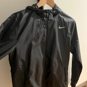 Svart vindjacka från Nike - Snygg svart vindjacka från Nike med dragkedja och huva. Perfekt för blåsiga dagar. Jackan har en enkel och stilren design med Nike-loggan på bröstet.
