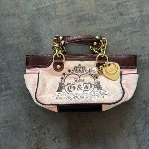 Äkta vintage handväska från Juicy Couture - Säljer en snygg rosa och brun handväska från Juicy Couture med guldfärgade kedjehandtag och dekorativa detaljer. Väskan har ett tryck med texten 'Love' och 'G&P' samt en hjärtformad detalj med bokstaven 'J'. Insidan är mönstrad med rosa kronor och texten 'Juicy'. Perfekt för att ge din outfit en lyxig touch!