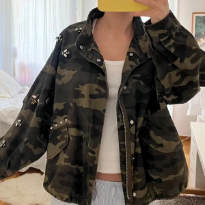 Camouflagejacka med pärldetaljer - Säljer en cool camouflagejacka med unika pärldetaljer. Jackan har en avslappnad passform med dragkedja och knappar framtill. Den har stora fickor och justerbar midja med snörning. Perfekt till våren!! Storlek M, sitter oversized. Den är i väldigt bra skick, inga tecken på användning alls.