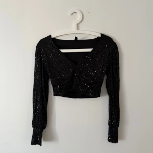 Svart glittrig croppad topp - Snygg svart glittrig topp med v-ringning och långa ärmar. Den är croppad och perfekt för en festlig kväll. Toppen har en elegant och stilren design som drar blickarna till sig.