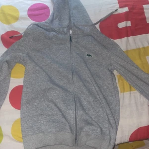 Grå hoodie från Lacoste - Säljer en stilren grå hoodie från Lacoste med dragkedja och den klassiska krokodilloggan på bröstet. Perfekt för en avslappnad look. Hoodien har långa ärmar och en bekväm passform. Säljer för att den är för liten storlek m