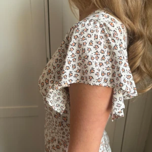 Blommig klänning med kort ärm - Säljer en söt blommig klänning med korta ärmar och v-ringning. Klänningen har ett fint mönster med små blommor i orange och svart på en vit bakgrund. Perfekt för sommarens varma dagar! Storlek 36/S