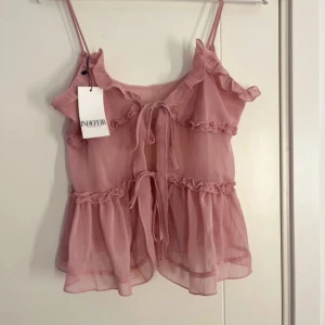 Rosa volanglinnetopp från Indefin - Säljer en söt rosa linnetopp från Indefin med volanger och tunna axelband. Toppen har en luftig design med knytning framtill och flera lager av volanger som ger en romantisk känsla.