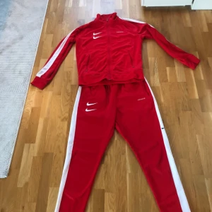 Röd träningsdräkt från Nike - Säljer en röd träningsdräkt från Nike med vita detaljer. Jackan har dragkedja och långa ärmar med vita ränder längs sidorna. Byxorna har en matchande design med vita ränder. Perfekt för träning eller en sportig look.