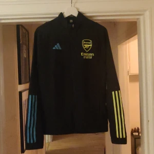 Arsenal Svart träningsjacka från Adidas - Säljer en svart träningsjacka från Adidas med Arsenal-logga och Emirates-tryck i gult. Jackan har långa ärmar med blå och gula ränder. Perfekt för fotbollsfans och träning. Använd en gång är som ny OBS FINNS PÅ FLER SIDOR  önskas fler bilder så är det bara att skriva 