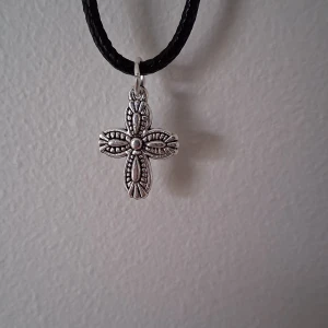 Silverfärgat korshalsband - Snyggt halsband med ett detaljerat korshänge i silverfärg. Hänget har ett intrikat mönster och hänger på en svart flätad rem. Perfekt för att ge en stilfull touch till din outfit.