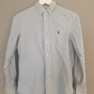 Blå och vit randig skjorta från Ralph Lauren - Säljer en stilren blå och vit randig skjorta från Ralph Lauren. Skjortan har en slim fit passform och är långärmad med knappar framtill. Perfekt för en klassisk look!