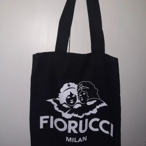 Svart shoppingväska från Fiorucci - Snygg svart shoppingväska från Fiorucci med ett ikoniskt tryck av två änglar och texten 'Fiorucci Milan' i vitt. Tillverkad i 100% bomull, perfekt för att bära med sig det nödvändigaste. Enkel och stilren design som passar till många tillfällen.