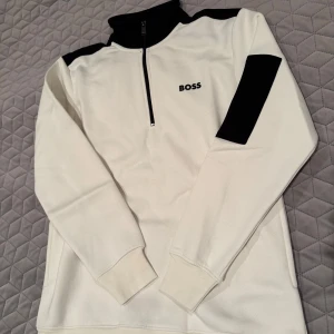 Vit tröja med dragkedja från Boss - Snygg vit tröja från Boss med svarta detaljer och dragkedja vid halsen. Tröjan har långa ärmar och en diskret logga på bröstet. Perfekt för en stilren look.
