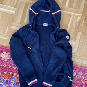 Mörkblå hoodie från Moncler - Säljer en snygg mörkblå hoodie från Moncler med dragkedja och huva. Den har ribbade muddar och ett Moncler-märke på ärmen. Perfekt för en casual look.