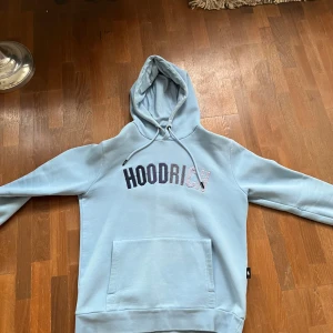 Ljusblå hoodie från Hoodrich - Säljer en snygg ljusblå hoodie från Hoodrich med tryck på bröstet. Den har en klassisk känguruficka och justerbar huva med dragsko. Perfekt för en avslappnad stil.