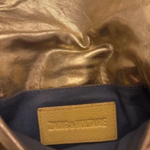 Guldig axelväska från Zadig & Voltaire - Snygg guldig axelväska från Zadig & Voltaire med kedjerem och dragkedja på baksidan. Perfekt för att ge en glittrig touch till din outfit. Kommer med en praktisk dustbag för förvaring. Nypris: 2355kr