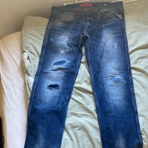 Blå jeans från Replay - Säljer ett par blå Replay jeans med slitna detaljer och hål för en cool look. De har en klassisk femficksdesign och knappgylf. Perfekta för en avslappnad stil.