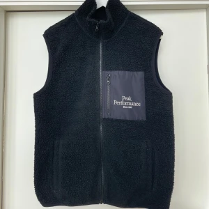 Svart fleeceväst/teddy från Peak Performance - I fint skick, teddymaterial, storlek L. Säljer en svart fleeceväst från Peak Performance med dragkedja och en praktisk bröstficka. Perfekt för lager-på-lager under kyliga dagar. Västen har en stilren design med märkets logga på bröstet.