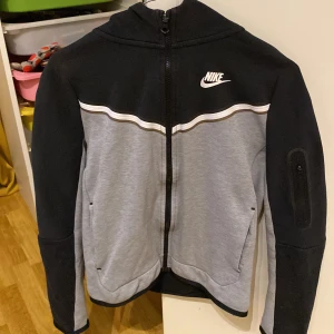 Svart och grå hoodie från Nike - Säljer en snygg svart vit och grå Nike tech med dragkedja och huva. Nike techen har en vit Nike-logga på bröstet och en cool design med vita ränder över bröstet. Perfekt för en sportig look!