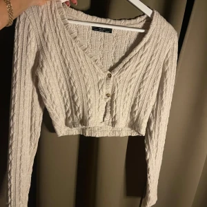 Beige stickad croppad kofta - Säljer en beige stickad kofta med v-ringning och knappar framtill. Den är croppad och har ett snyggt kabelstickat mönster. Perfekt för lager-på-lager-stil! Säljs inte längre!